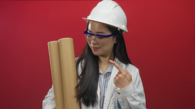 Young chinese woman architect in hardhat holds blueprints against red background, wearing safety goggles and white coat.