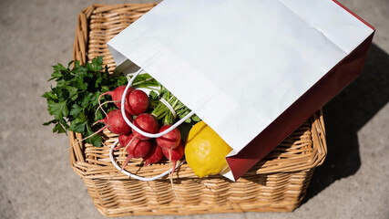 Fresh produce in a woven basket with a paper bag on a table in bright daylight