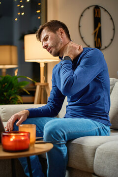 Man sitting on sofa and holding his neck, suffering from pain. Person with discomfort and muscle tension due poor posture during work at laptop