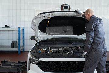 Middle aged Caucasian man inspecting engine compartment of modern car in auto repair shop, standing beside open hood and wearing work gloves, focusing on vehicle maintenance