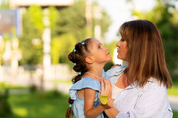 In the soft glow of late afternoon, little girl in blue-striped dress looks up intently at her mother, who leans in with pursed lips, as they hold single leaf between them in leafy park setting