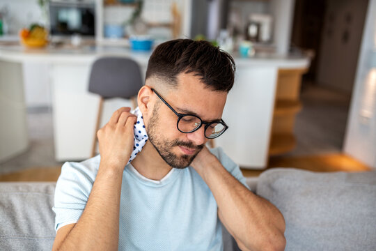 Young man sitting on a sofa and using an ice pack for neck pain and cervical spine relief while suffering from muscle tension or a stiff neck and stress from poor posture at home