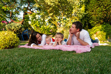 Parents and two kids lie on a fluffy pink throw in the grass, chatting and relaxing among trees and shrubs during golden hour