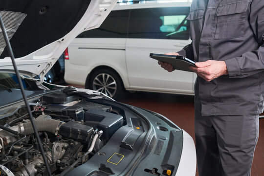Unrecognizable man inspecting car engine while holding digital tablet in auto repair shop, standing next to open hood and focusing on diagnostic process, vehicle visible in background
