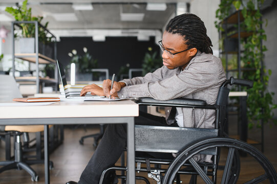 Focused african american employee using laptop and taking notes in wheelchair at modern office