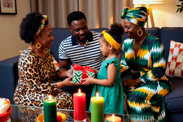 Obraz premium Black family gathering in living room, middle aged women and man smiling while young girl receiving wrapped gifts, colorful candles on table, festive celebration atmosphere