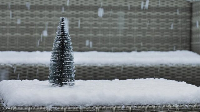 Small artificial pine tree sitting on a snow-covered outdoor bench during a gentle winter snowfall.