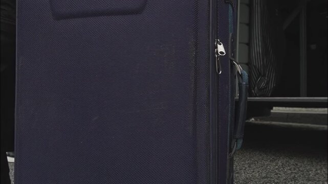 Person rolls dark blue suitcase with zipper and part of handle. Plain texture and design of luggage suggest ready for travel and storage