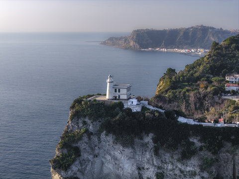 Aerial view of a stark white lighthouse perched atop a rugged cliff overlooking the serene blue sea against the backdrop of distant hills, Capo Miseno, Bacoli, Campania, Italy.