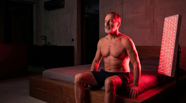 Fit middle‑aged man sitting on bed in dark bedroom, illuminated by large red light therapy panel. Recovery, performance, biohacking and modern wellness rituals.