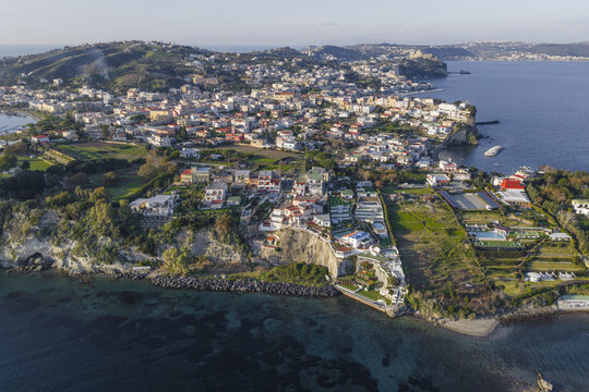 Aerial view of a coastal town with colorful buildings nestled against the azure sea, cliffs, and lush greenery, Capo Miseno, Bacoli, Campania, Italy.