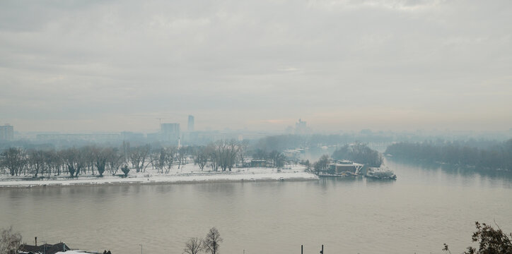 Winter river panorama of Belgrade with snowy riverbank, bare trees and misty city skyline, creating calm atmospheric urban landscape in cold season.
