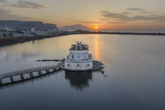 Aerial view of Casina Vanvitelliana standing gracefully on Fusaro Lake under a sky painted with the warm hues of the setting sun, Bacoli, Campania, Italy.
