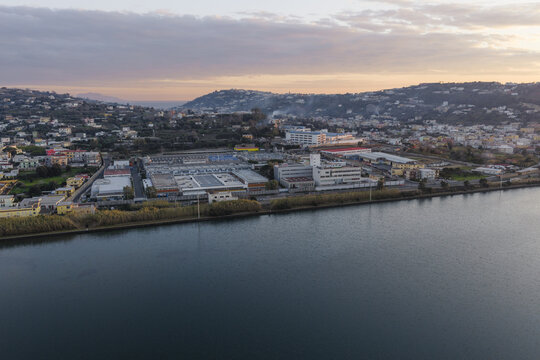 Aerial view of a sprawling industrial zone meets the tranquil waters of a lake, nestled amidst rolling hills under a soft, pastel sky, Fusaro Lake, Bacoli, Campania, Italy.
