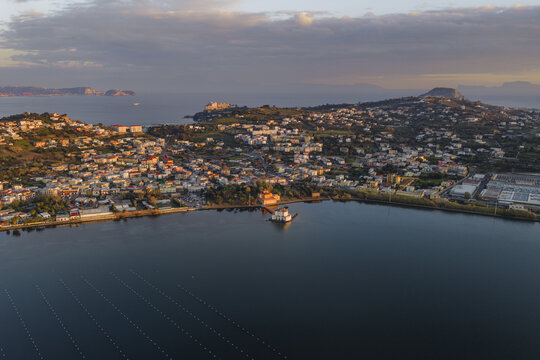 Aerial view of a tranquil lake mirroring the sky, with quaint buildings nestled along the shore and distant hills under a soft, golden light, Fusaro Lake, Bacoli, Italy.