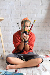 A young artist in an orange beanie and shirt sits cross-legged, playfully hiding behind a bundle of paintbrushes against a white brick wall in their creative studio.
