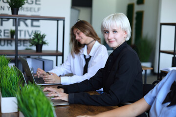 A confident young woman with platinum hair smiles at the camera while working on a laptop in a modern, plant-filled office. Colleagues focus nearby, creating a vibrant