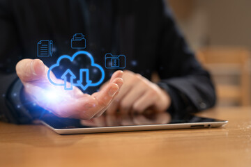 Close-up of a hand interacting with cloud computing icons above a digital tablet, representing data storage, file upload and download, cloud technology, digital transformation.