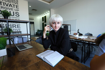 A confident student with platinum hair sits at a modern desk, pen in hand, smiling softly at the camera. Open textbook and laptop nearby, motivational wall quotes inspire focus in this bright