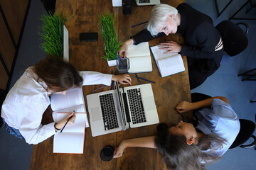 An overhead view captures three students studying together at a wooden table, surrounded by laptops, notebooks, and green plants. Their focused and relaxed postures 