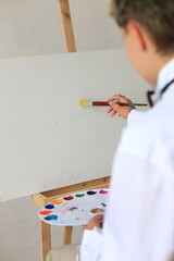 A young artist in a white shirt focuses intently, applying yellow paint to a blank canvas on an easel, beginning a new artwork with a colorful palette nearby.