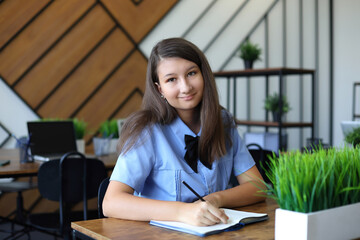 A cheerful student in a light blue uniform smiles softly at the camera while writing in a notebook. She sits at a wooden desk surrounded by green plants and modern decor