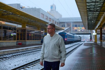 A man in a light jacket stands alone on a snow-dusted train platform as gentle flakes fall. A blue train is visible in the distance, and the station's 