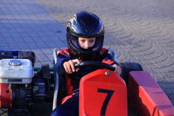 A young Caucasian boy in a black helmet drives a go-kart with a red number 7 on it. The background features a paved surface, indicating a racing environment.