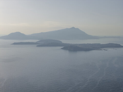 Aerial view of islands resting tranquilly on the calm, silvery water, with distant mountains shrouded in a soft, hazy atmosphere, Procida, Campania, Italy.