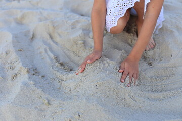 A child in a white dress crouches on a sandy beach, her hands gently shaping the soft sand into patterns, capturing a moment of simple, tactile play.