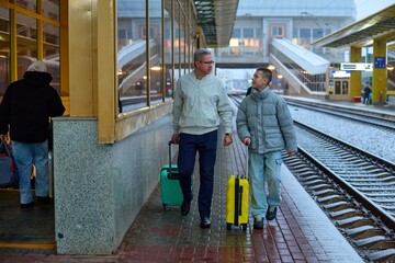 A father and son walk together along a snowy train platform, each pulling a brightly colored suitcase. They share a moment of connection as they head 