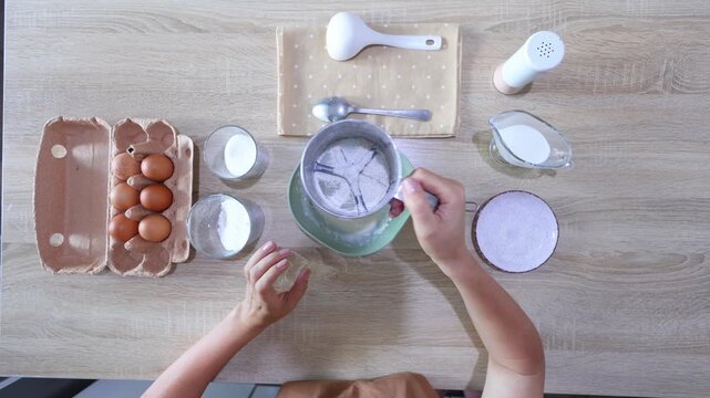 A top-down view shows hands sifting flour into a bowl, then whisking it smoothly. Surrounding ingredients eggs, milk, sugar, and utensils suggest the start of a baking recipe. Clean, minimalist setup 