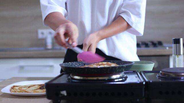 A person expertly flips a golden-brown pancake in a skillet using a flick of the wrist, then slides it onto a plate with other finished pancakes, ready to serve.