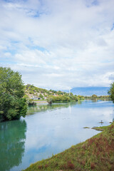 Fototapeta premium Calm river flowing through a lush green landscape with trees, grassy banks, and distant hills under a soft cloudy sky. Reflections on the water create a tranquil atmosphere, while birds resting