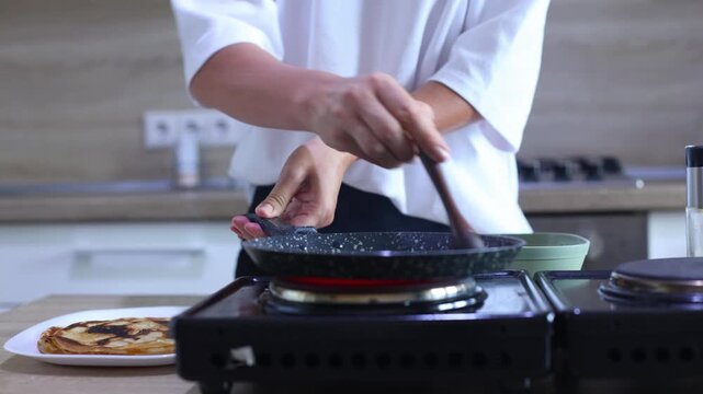 A person prepares to cook another pancake by greasing a hot skillet with oil, then pouring in batter from a bowl and swirling it to form a perfect circle.