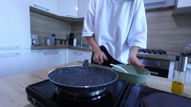 A person prepares to cook another pancake by greasing a hot skillet with oil, then pouring in batter from a bowl and swirling it to form a perfect circle.