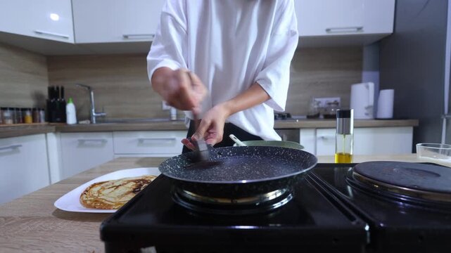 A person prepares to cook another pancake by greasing a hot skillet with oil, then pouring in batter from a bowl and swirling it to form a perfect circle.