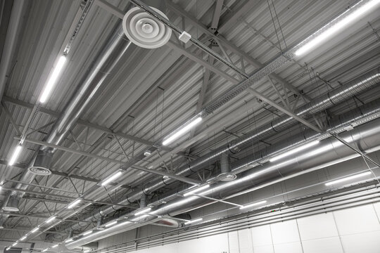 Wide view of an industrial ceiling featuring exposed HVAC ducts, ventilation vents, and suspended LED lighting in a large, clean workspace. Includes structural beams and copy space