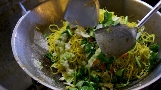 stir with large spoons in a wok topview a plate of stir fried yellow noodle with lots of saag veges.