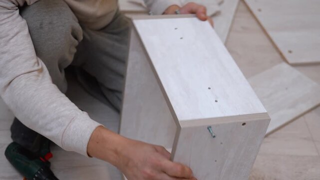 Close-up of hands of man assembling new wooden furniture at home using a cordless screwdriver screwdriver to fasten particle board panels during home renovation by himself