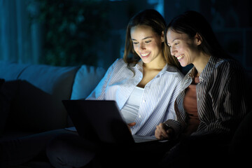 Two happy friends watching media online on laptop in the night