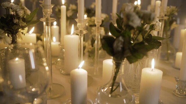 Elegant table setting with numerous burning white candles in glass candlesticks and vases with flowers creating a romantic atmosphere
