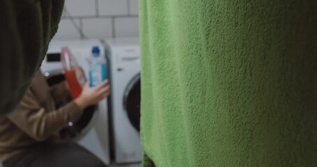 A man kneels in front of a washing machine holding detergent, unsure which one to use. Concept of everyday decision-making, household chores, confusion, and domestic routine.