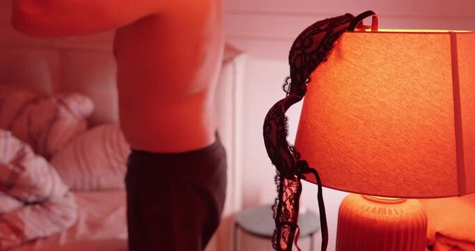 A dimly lit bedroom scene with lingerie draped over a bedside lamp while a person moves in the background. Concept of intimacy, temptation, secrecy, and relationship tension.