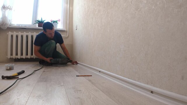 Male worker doing home renovation, professionally installing white plastic skirting board on the wall, using a power drill and hammer for finishing touches on the new laminate flooring