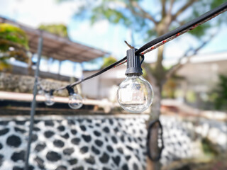 Obraz premium Close-up of outdoor string lights hanging in a backyard patio with a blurry garden background on a sunny day. Ideal for cafe, event, or home decor lifestyle concepts