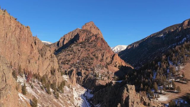 A cinematic aerial vista highlights a sharp mountain peak rising above steep rocky canyon walls. Dramatic deep shadows and winter snow showcase the immense scale of this rugged mountain environment.