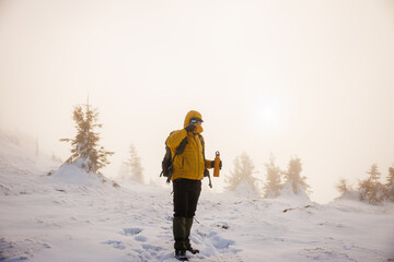 Man hiker in yellow winter jacket holding thermos on snowy mountain peak during sunset. Refreshment by hot drink during hiking in mountains
