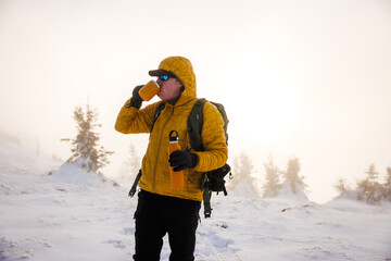 Man hiker in yellow jacket holding thermos and drinking hot tea from mug at mountain sunset. Refreshment during winter trekking in cold misty weather