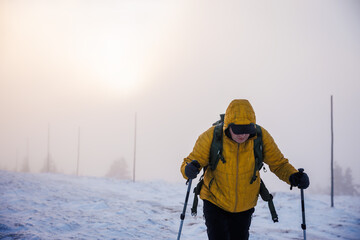 Winter hiking. Active man in yellow jacket with hiking poles climbing at mountain peak in misty cold weather. Challenging winter trek in fog and snow